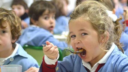 Una niña, en un comedor escolar, se lleva la comida a la boca.