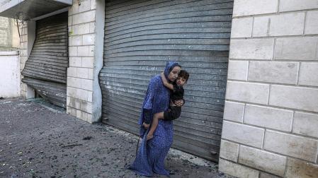 Una mujer aúpa a un niño descalzo entre los cristales de ventanas rotos por los bombardeos en la ciudad de Gaza hace dos semanas
