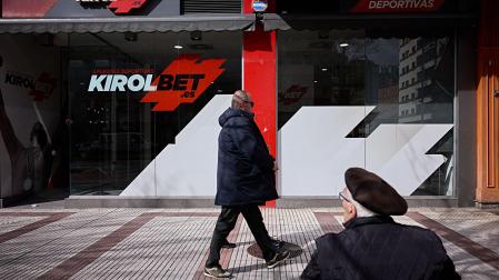 Dos hombres caminan  y uno descansa en un banco delante de un local de apuestas ubicado en Pamplona
