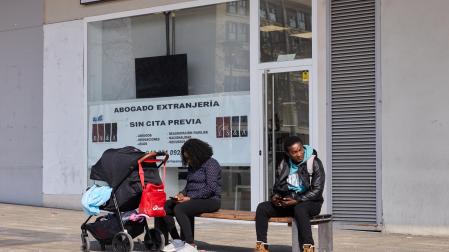 Dos personas consultan su móvil en las inmediaciones de la oficina de Extranjería, en el barrio de Buztintxuri. De fondo, un cartel publicita los servicios de un abogado.