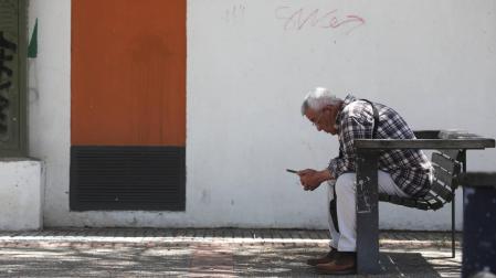 Un jubilado observa su móvil sentado en un banco a la sombra