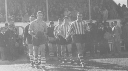 Martino y Zarra, capitanes de la Peña Sport y el Atlético de Bilbao, encabezan la salida al terreno de juego. Detrás, la grada poblada