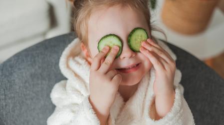 Adorable little girl 3 years old in a soft home dressing gown with cucumbers in her hands plays in the beauty salon at home. The child makes beauty treatments from natural items.