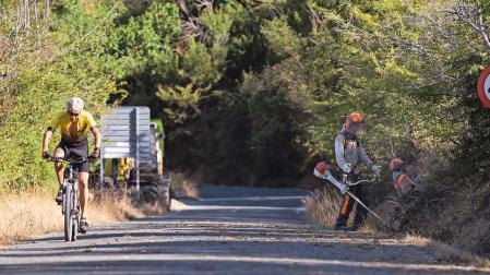Labores de desbroce, ayer, junto a la carretera de acceso al Fuerte mientras uno de los ciclistas circula por la vía.