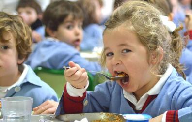 Una niña, en un comedor escolar, se lleva la comida a la boca.