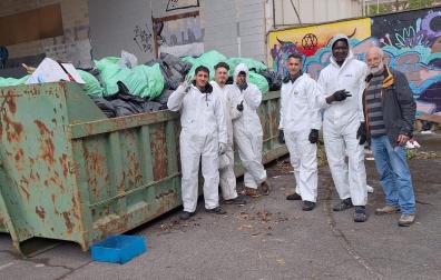 Jóvenes sin hogar introducen en un contenedor la basura recogida del interior del edificio abandonado de la antigua ikastola Jaso en Echavacoiz, donde lvan meses sobreviviendo. A la derecha, Mikel Otazu, profesor jubilado y voluntario de una de las entidades sociales que colaboran en la recogida.