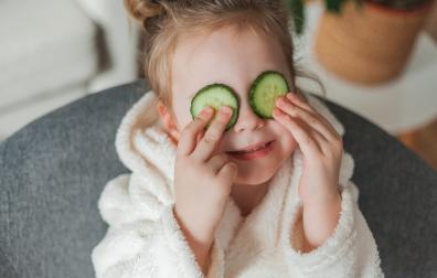 Adorable little girl 3 years old in a soft home dressing gown with cucumbers in her hands plays in the beauty salon at home. The child makes beauty treatments from natural items.
