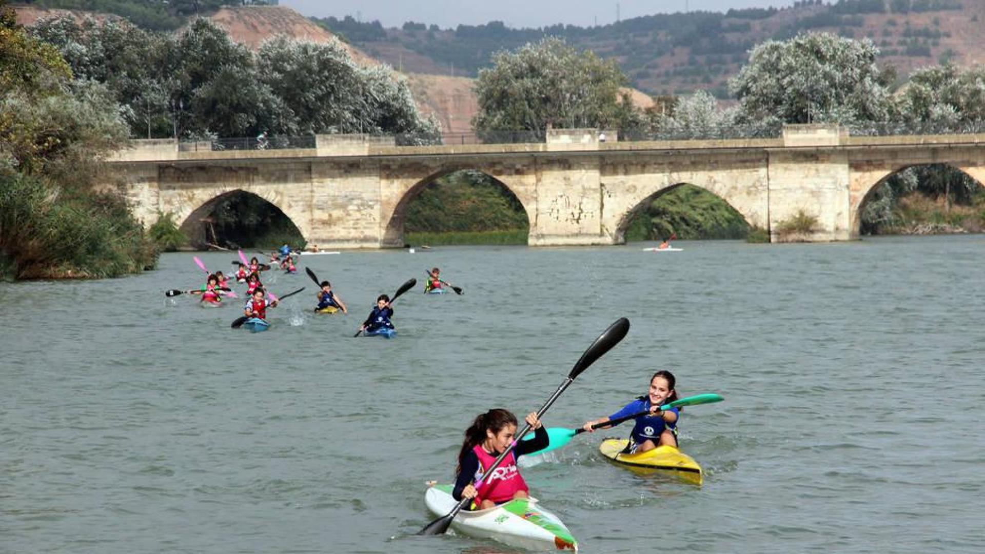 Varios jóvenes navegan con piraguas por el río Ebro con el puente de Tudela a sus espaldas.