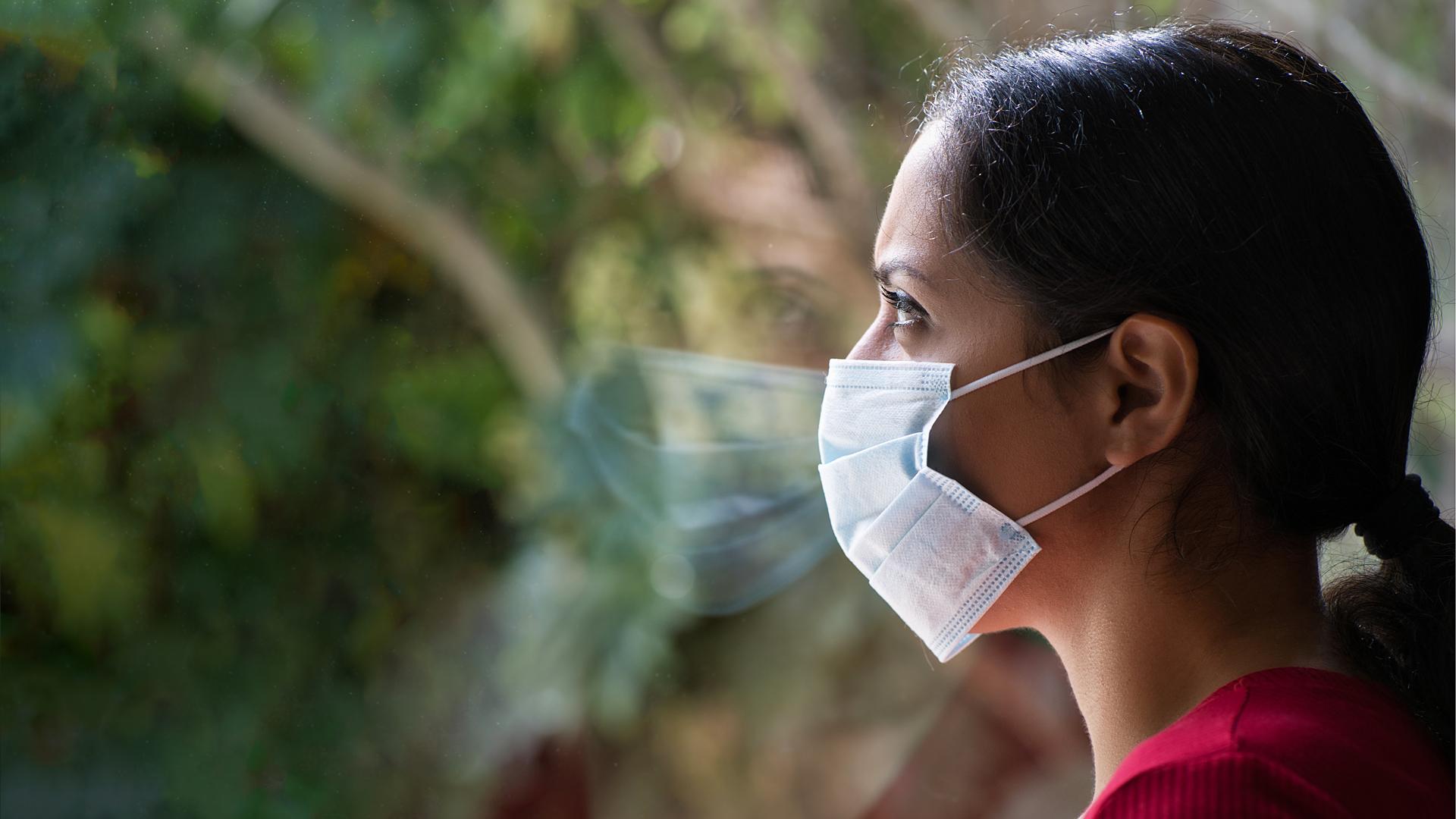 Una mujer protegida con una mascarilla observa a través de una ventana