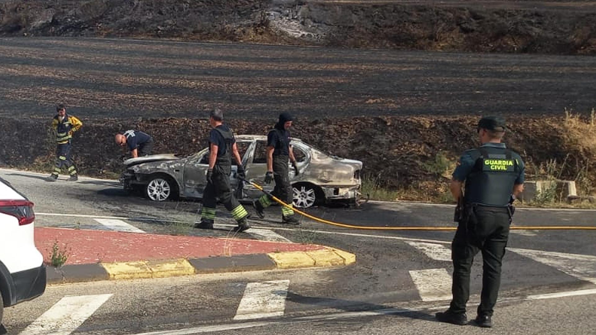 Un agente de la Guardia Civil junto a los bomberos y el vehículo incendiado