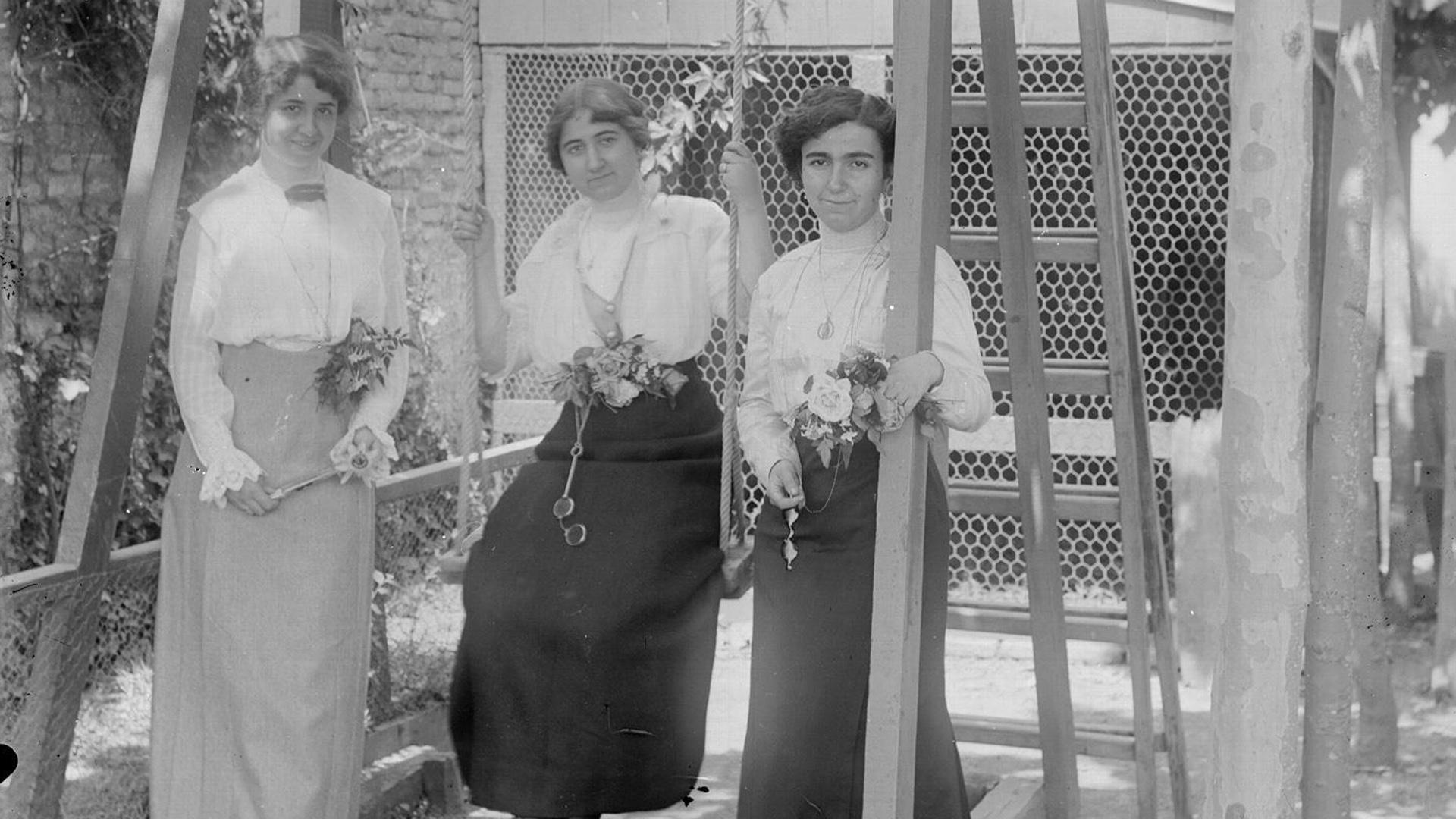 Retrato en plano entero de 3 mujeres con vestidos, ramos de flores y anteojos en un columpio instalado en la zona de juegos de "Garden House", la casa de recreo de la familia Altadill Aldave situada en el paraje de La Magdalena en Pamplona