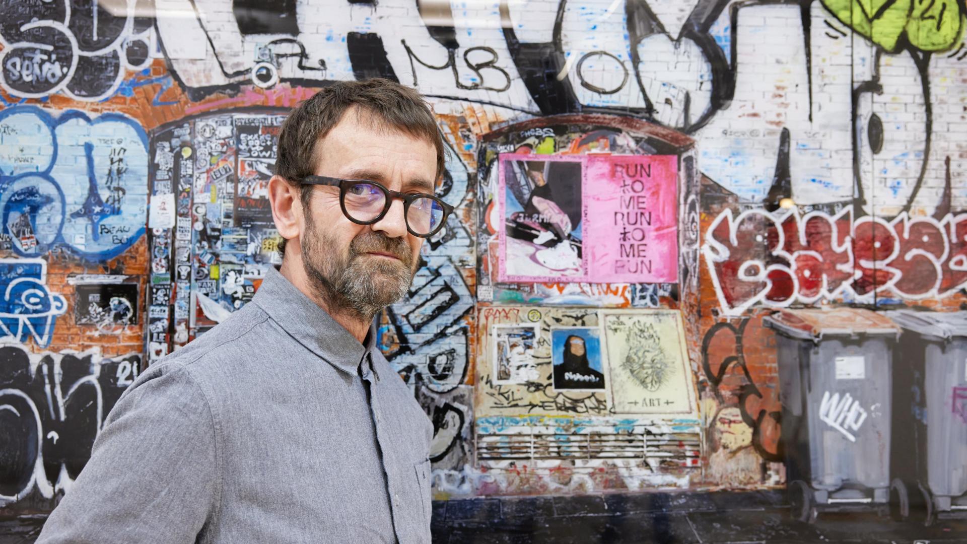 Pablo Ochoa de Olza posa junto a una pintura llena de grafitis y anuncios comerciales que refleja una pared pintada frente al Museo de Arte Contemporáneo de Barcelona.