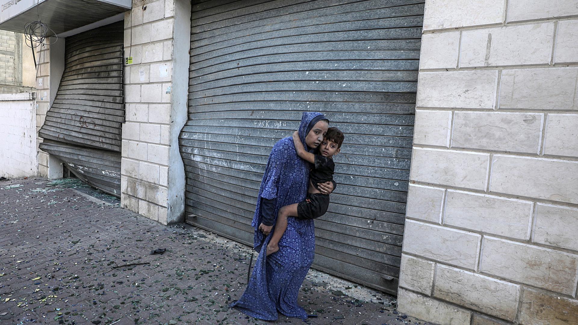 Una mujer aúpa a un niño descalzo entre los cristales de ventanas rotos por los bombardeos en la ciudad de Gaza hace dos semanas