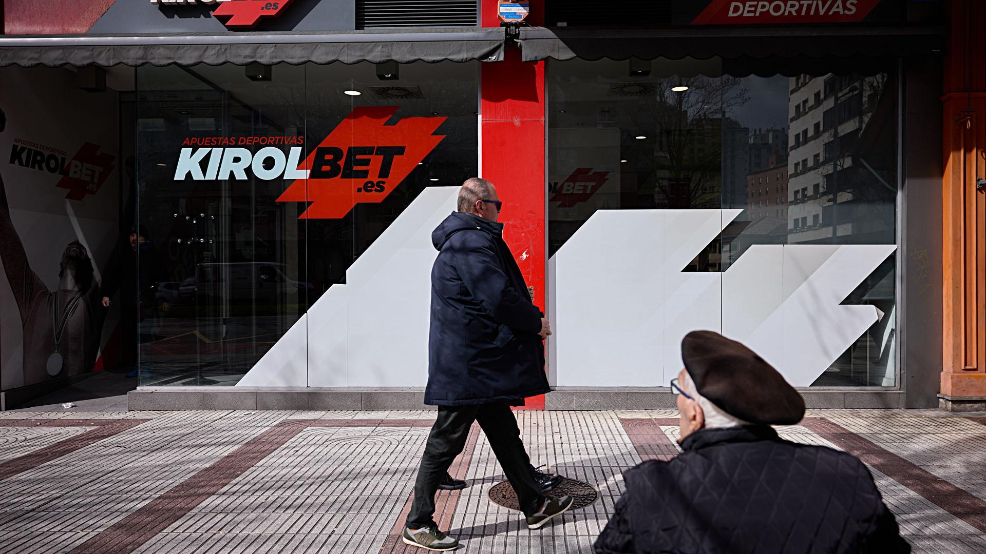 Dos hombres caminan  y uno descansa en un banco delante de un local de apuestas ubicado en Pamplona