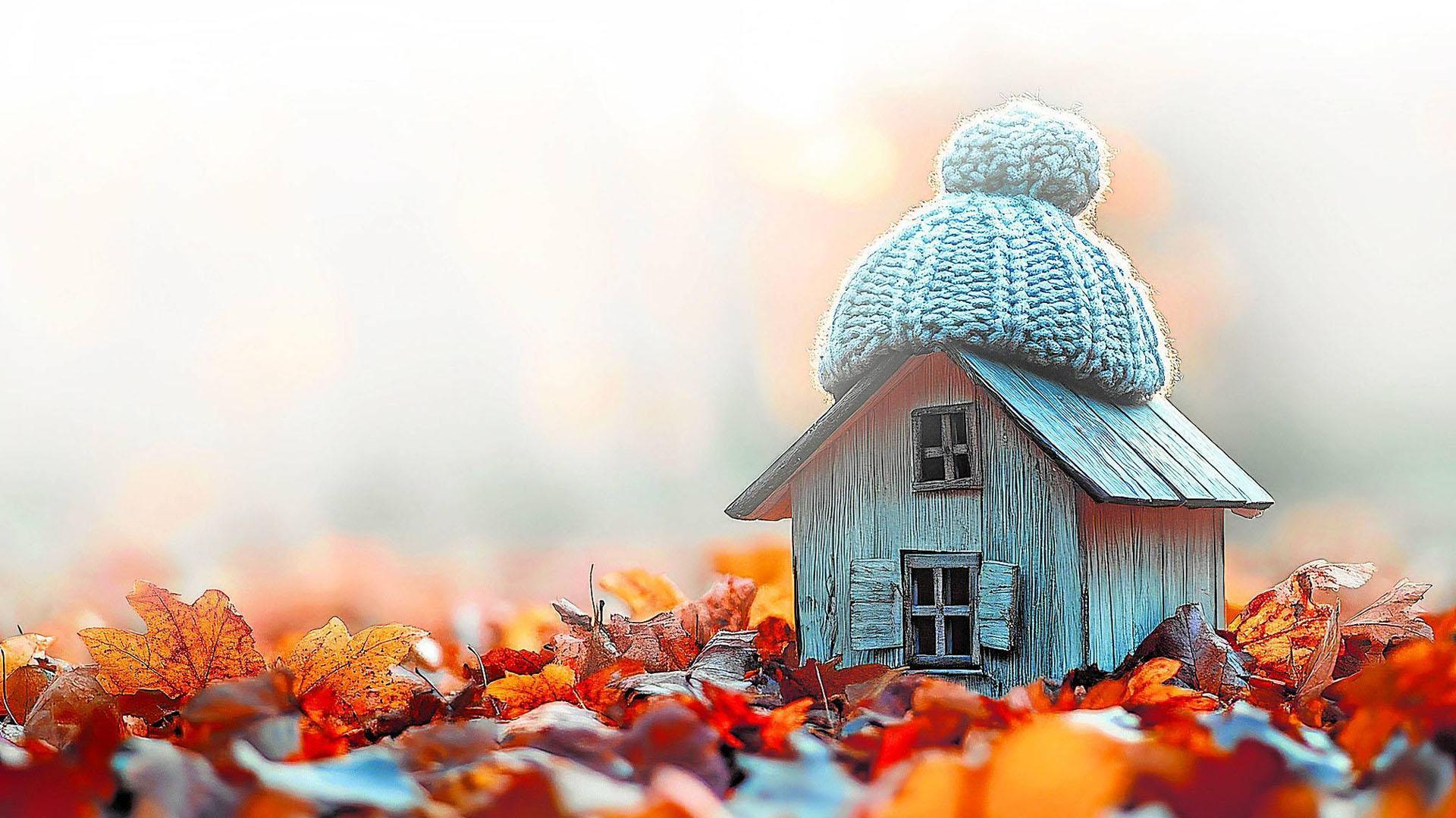 Una encantadora casa en miniatura cubierta cálidamente por un gorro de punto, enclavada entre coloridas hojas de otoño, lista para el invierno