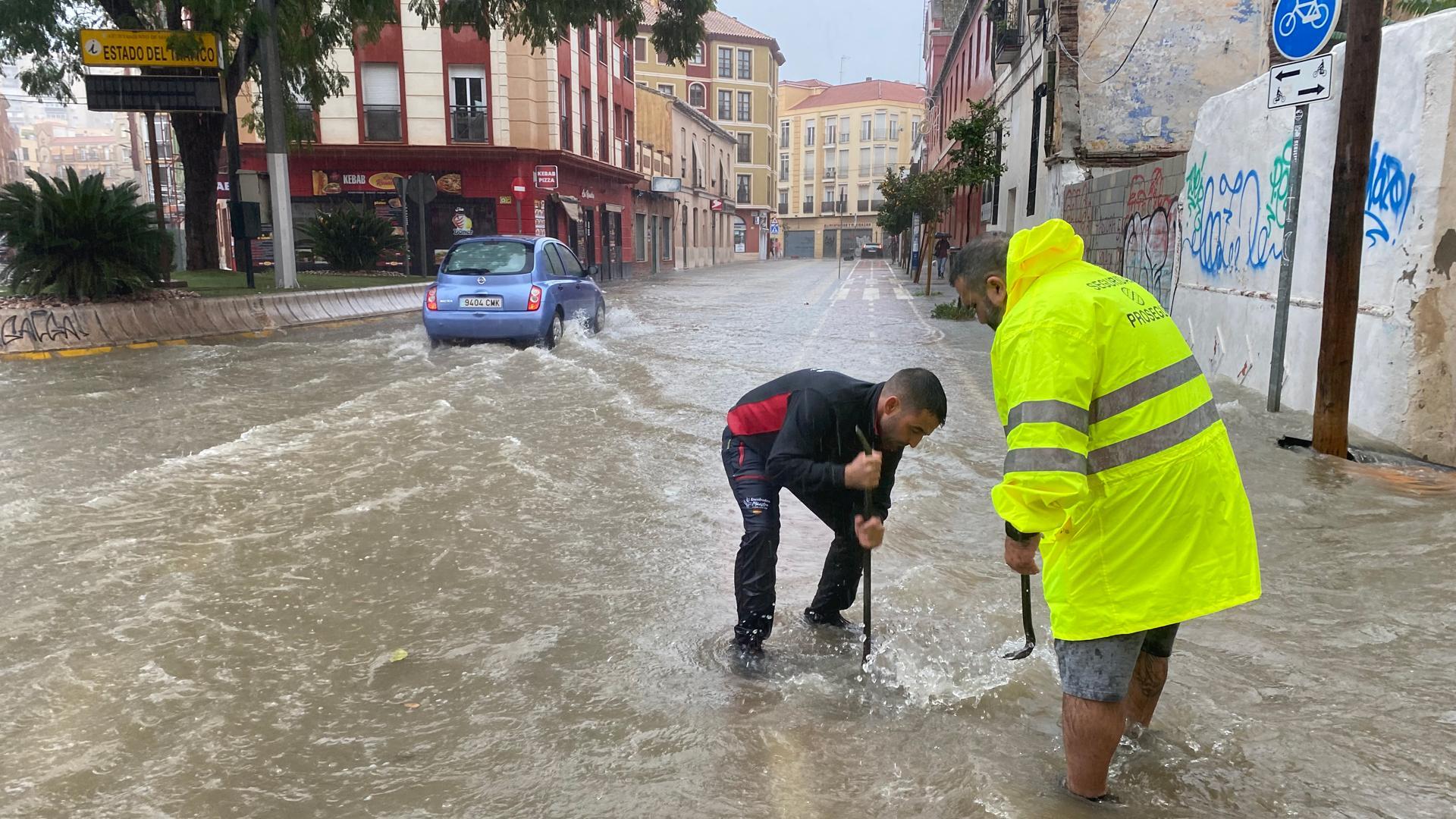 Varias personas intenta levantar la tapa de una alcantarilla en una calle inundada de agua debido a las fuertes lluvias y granizo que se han registrado este miércoles en Málaga