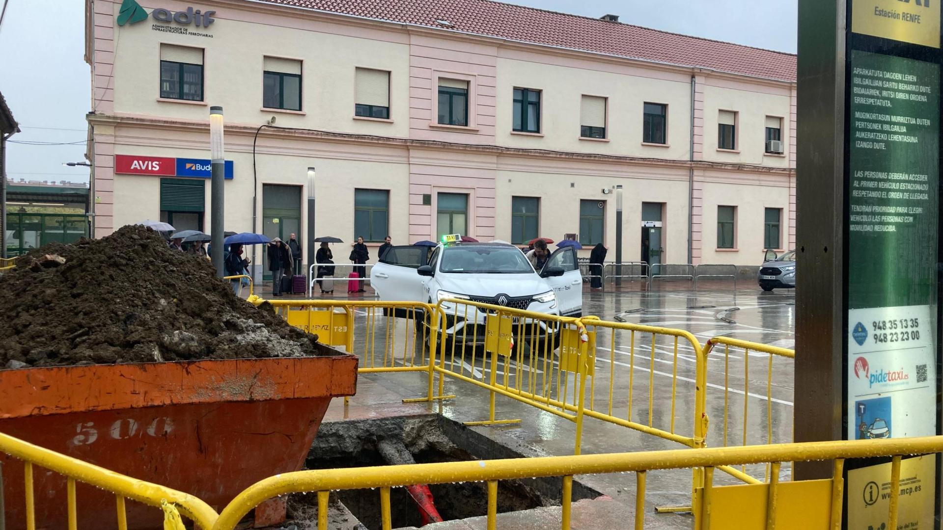 Una cola de pasajeros espera a un taxi bajo la lluvia en la estación de tren de Pamplona. En primer plano, las obras para construir una pérgola para resguardarse