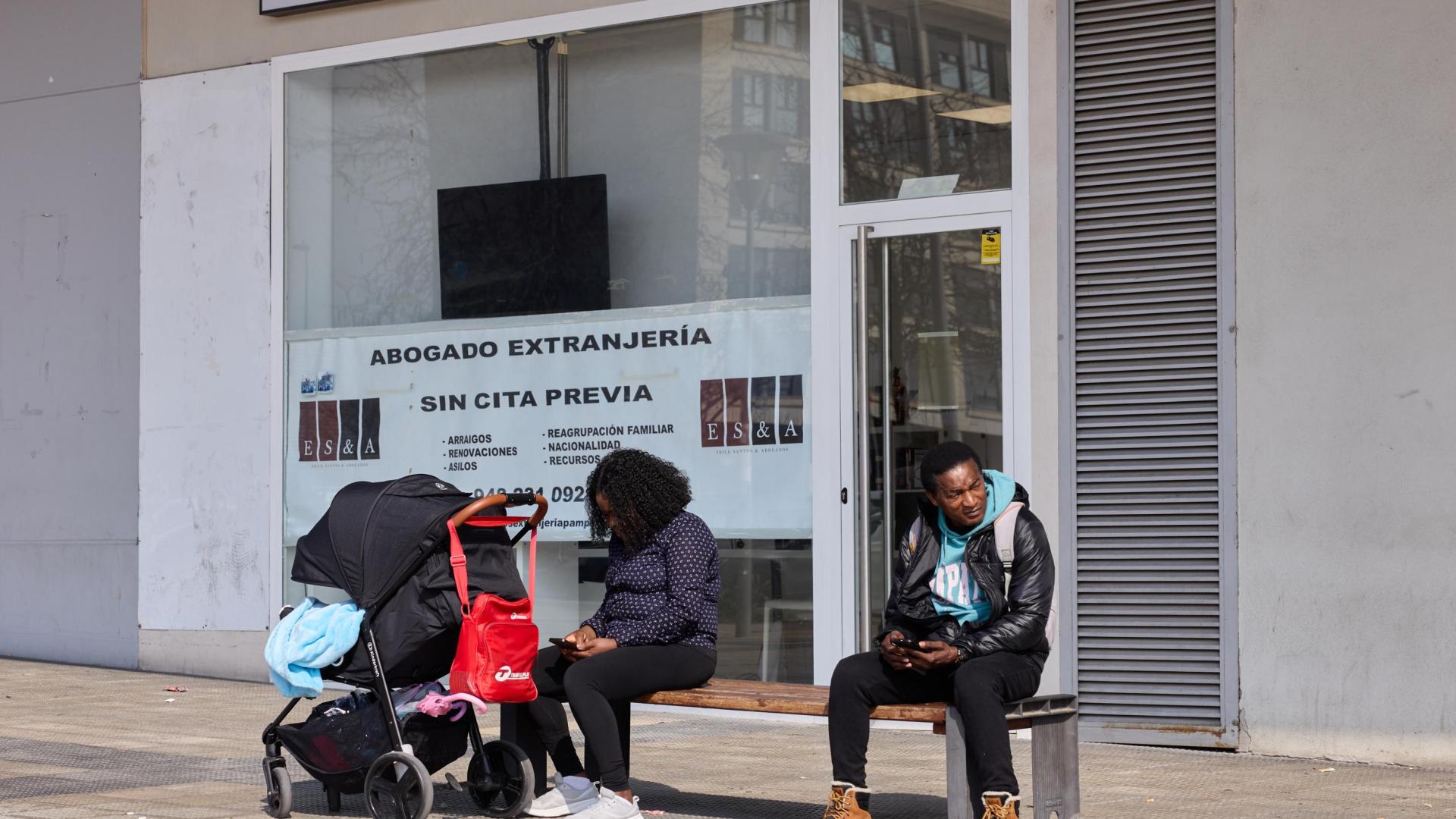 Dos personas consultan su móvil en las inmediaciones de la oficina de Extranjería, en el barrio de Buztintxuri. De fondo, un cartel publicita los servicios de un abogado.