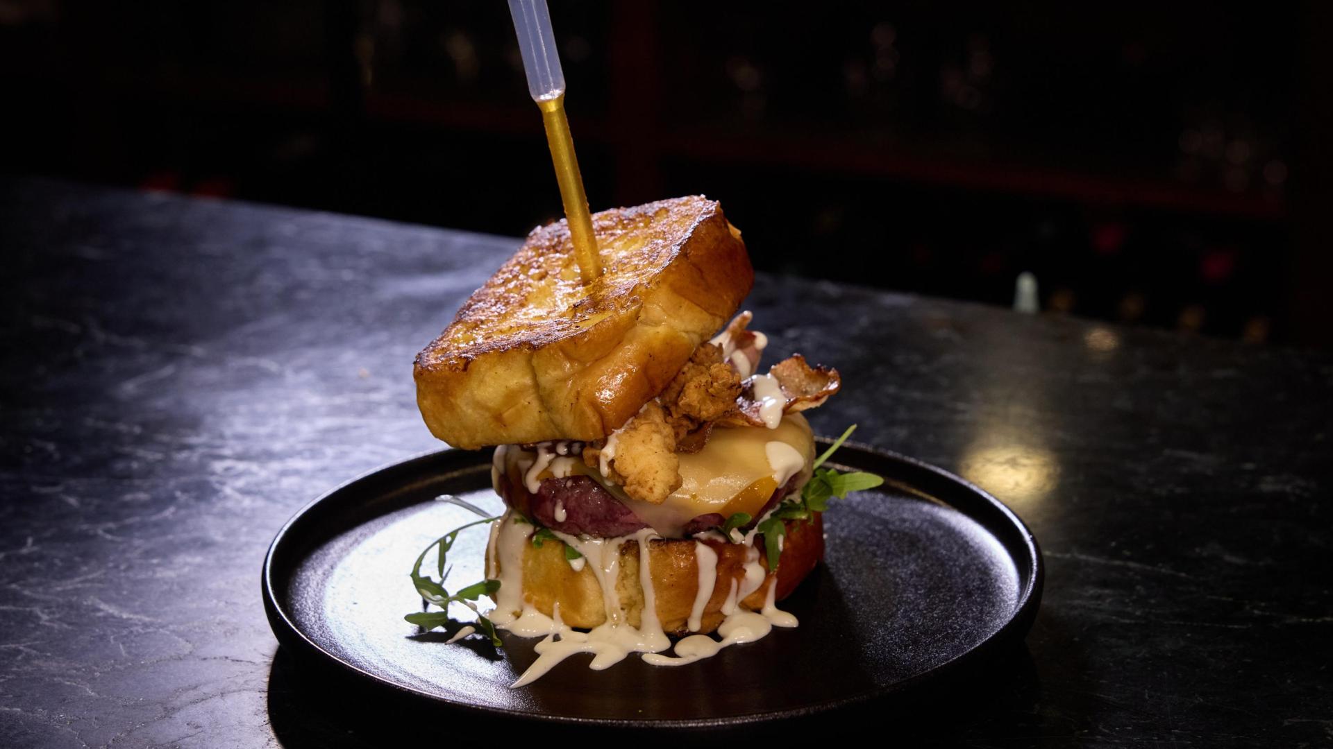 Idea del jefe de Gastro Okei Errialde con inspiración estadounidense, Fermín Team. En french toast, carne con doble de queso, rúcula, salsa ranchera con queso azul, bacon ahumado, pollo frito y salsa hot honey (miel picante).