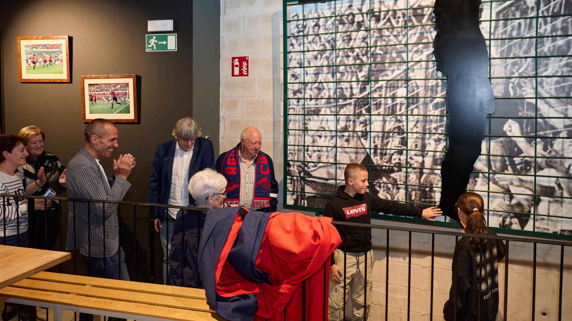 Oihan y Nikole, los hijos de Puñal, junto a la valla de El Sadar que subía Chiquiín y que fue ayer descubierta en  el Txoko de El Sadar, donde va a quedar expuesta. La silueta es la de Enrique Martín, que también aparece en la imagen junto a las tres hijas de Chiquilín, a la izquierda.