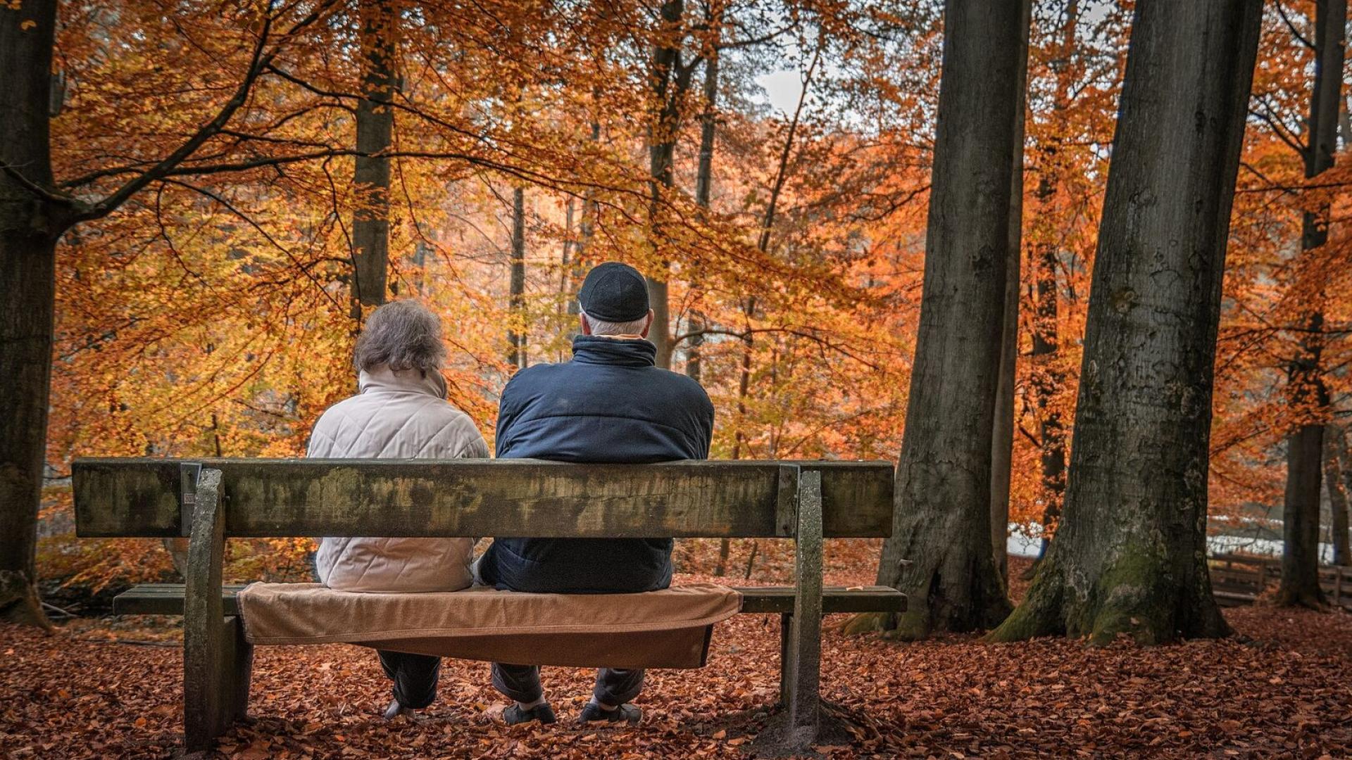 Dos personas jubilados, sentadas en el banco de un paseo en otoño.