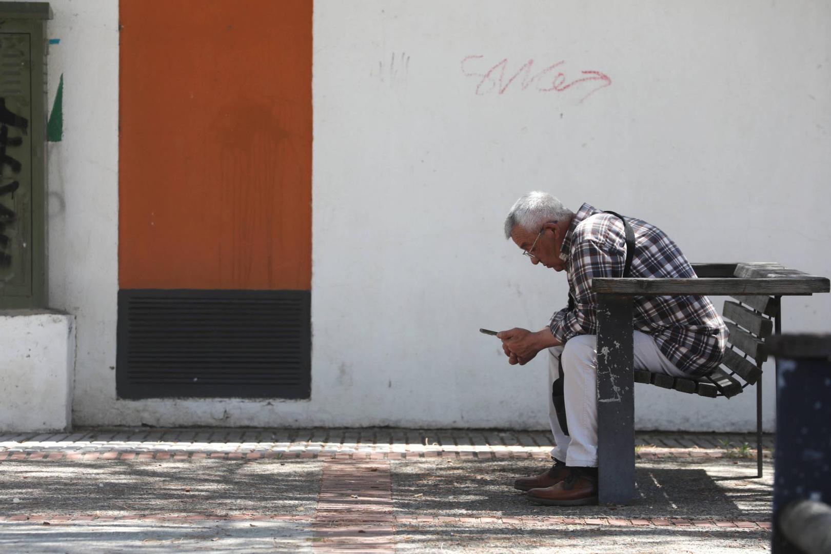 Un jubilado observa su móvil sentado en un banco a la sombra