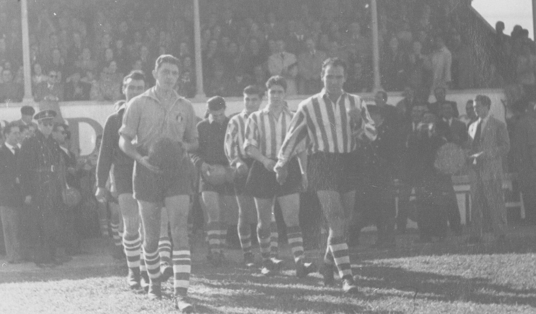 Martino y Zarra, capitanes de la Peña Sport y el Atlético de Bilbao, encabezan la salida al terreno de juego. Detrás, la grada poblada