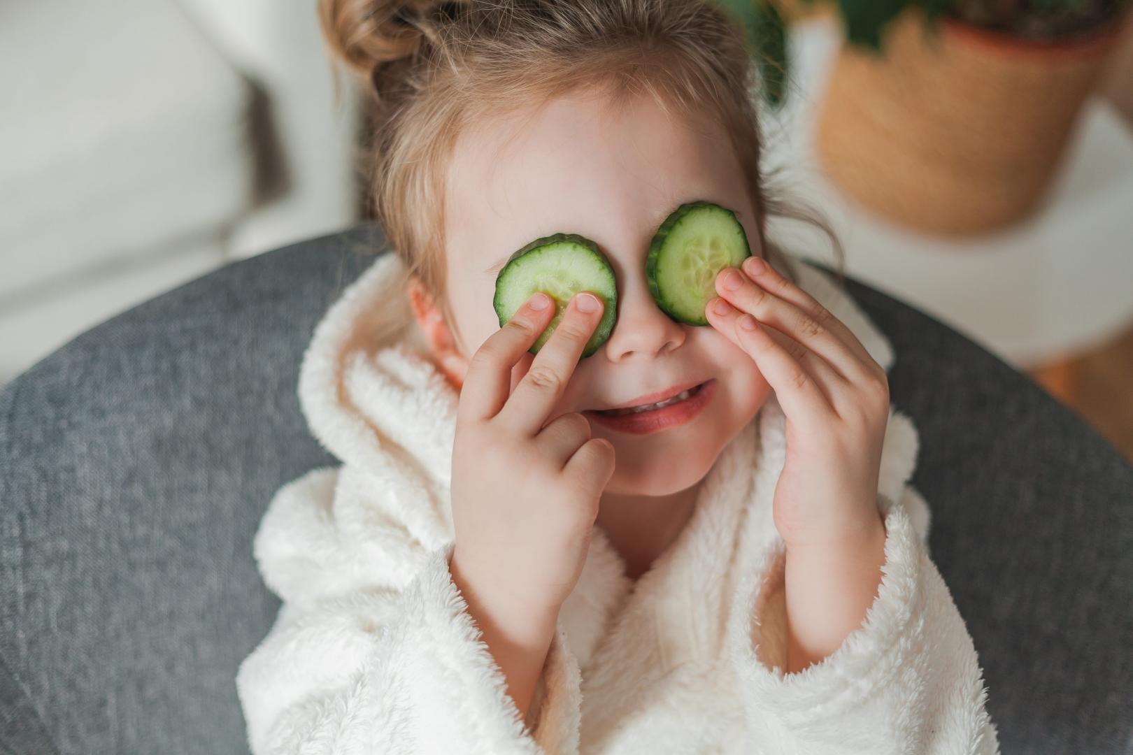 Adorable little girl 3 years old in a soft home dressing gown with cucumbers in her hands plays in the beauty salon at home. The child makes beauty treatments from natural items.