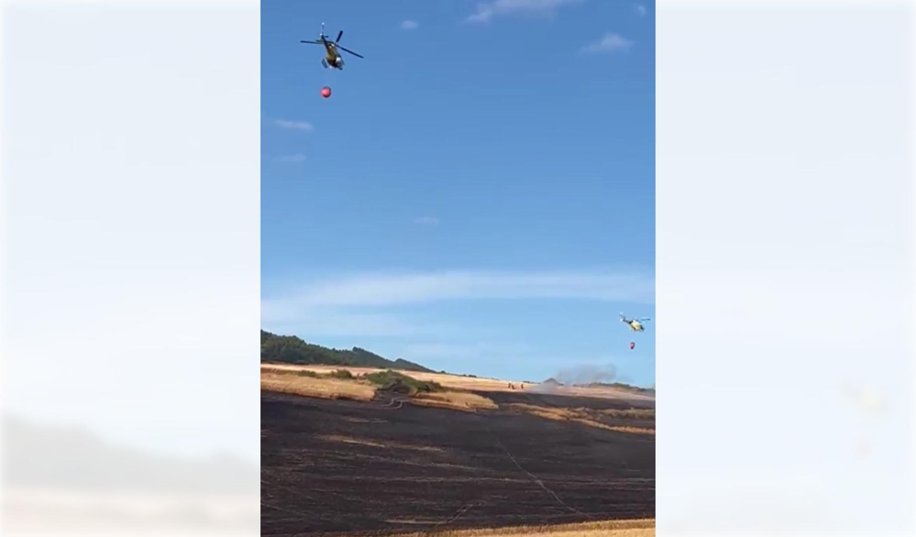 Los helicópteros recogen agua de la balsa de Ezkoriz y apagan las llamas del fuego en el campo de cereal cercano.