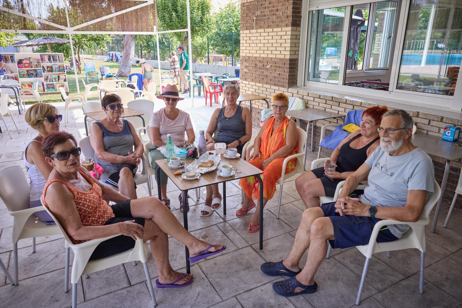 De izq. a dcha.: María Antonia Alfaro, Montse Boteargues, María Jesús Ábrego, Inma Bueno, Rufo Ayerbe, Mari Carmen del Río, María José González y Natxo Salinas desayunan en la piscina municipal de Miranda de Arga, con la biblioteca móvil al fondo.