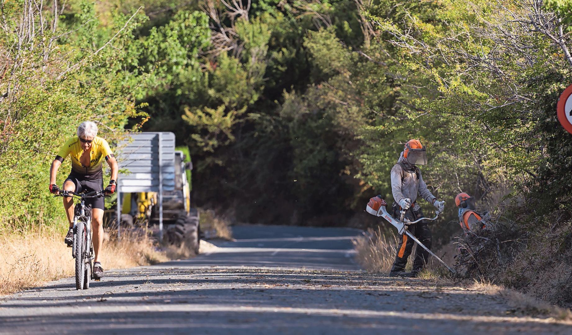 Labores de desbroce, ayer, junto a la carretera de acceso al Fuerte mientras uno de los ciclistas circula por la vía.