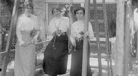 Retrato en plano entero de 3 mujeres con vestidos, ramos de flores y anteojos en un columpio instalado en la zona de juegos de Familia Altadill Aldave